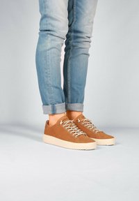 Zapatillas de ante marrón con suela de goma beige, diseño con cordones y detalles de costura. Combinadas con jeans azules enrollados.