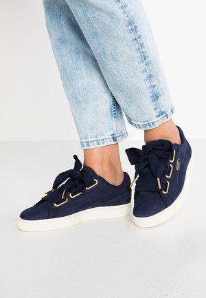 Personne portant un jean court bleu clair et des baskets bleu marine avec de grands nœuds en tissu et des semelles blanches, debout sur une surface blanche.
