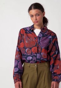 Košeľa s dlhým rukávom na zapínanie na gombíky, tmavomodrá so vzorom kvetov v červenej a fialovej farbe; doplnená o vysoký pás na olivovo zelených nohaviciach.