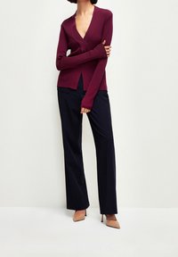 Cardigan en tricot bordeaux avec un col en V profond et des manches longues, doté de fentes latérales, associé à un pantalon bleu marine sur mesure et des talons nus.