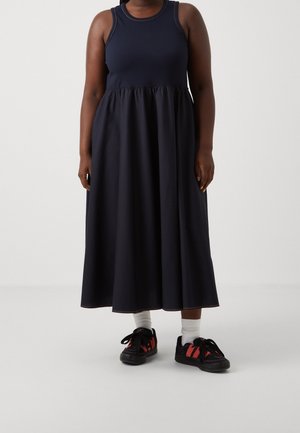 Personne portant une robe bleu marine sans manches avec une taille froncée, des chaussettes blanches et des baskets noires avec des rayures rouges, debout devant un fond uni.