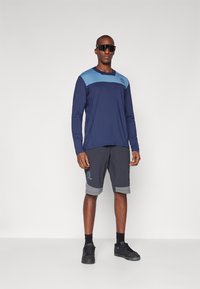 Langarmige marineblauwe shirt met lichtblauwe accenten en logo; grijze shorts met lichtgrijze manchetten en zijneten; zwarte sneakers en zonnebril.