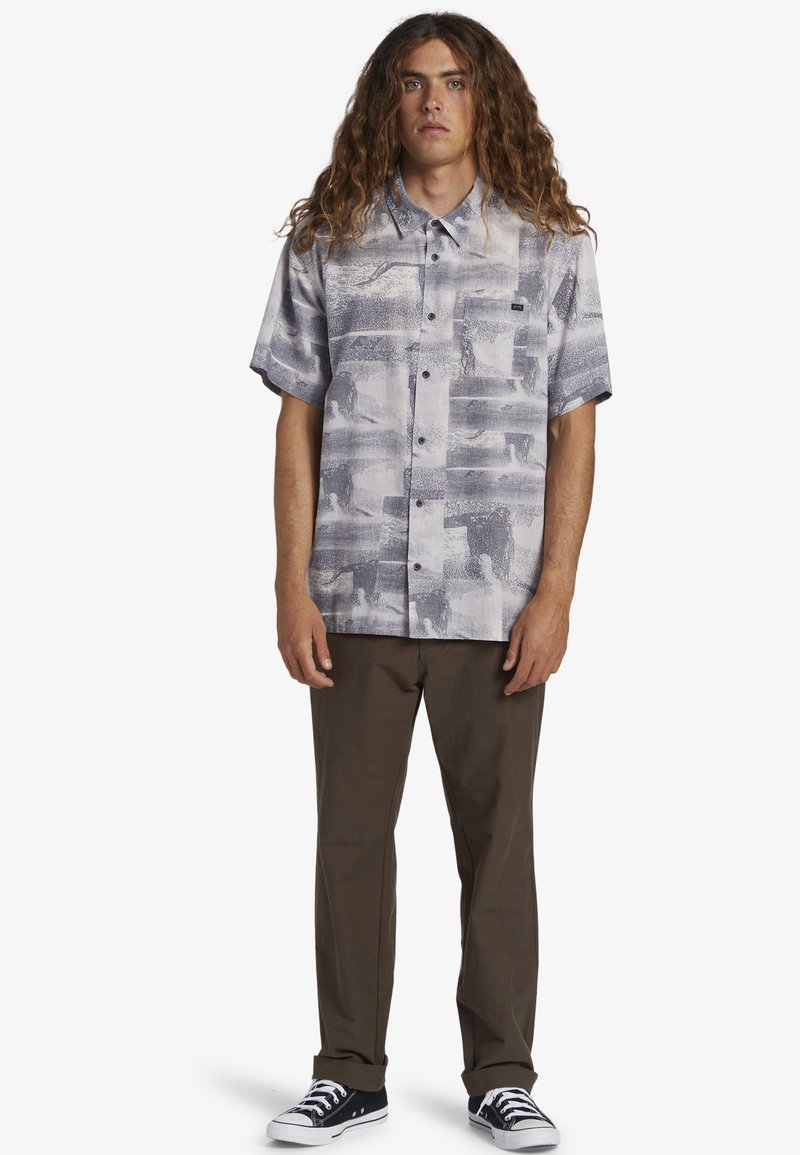 Camisa de botones de manga corta en gris claro con un patrón abstracto desvanecido, combinada con pantalones marrones y zapatillas negras.