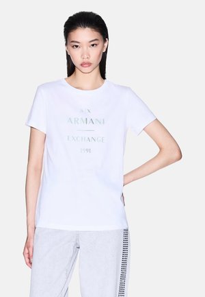 Giovane donna con capelli lisci e scuri indossa una t-shirt bianca Armani Exchange e pantaloni chiari con motivo a quadrati neri sul lato.