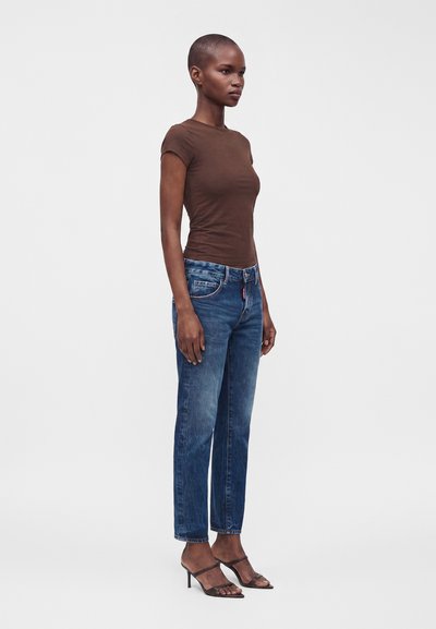 Femme debout de profil portant un t-shirt marron ajusté à manches courtes, un jean bleu et des sandales à talons noires à bouts ouverts sur un fond uni.