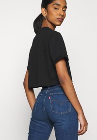 Schwarzes, kurzes T-Shirt kombiniert mit blauen, hochgeschnittenen Jeans, die ein rotes Etikett und Details mit abgerundeten Taschenstichen aufweisen, aus glattem Baumwollmaterial.
