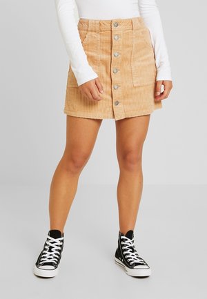 Femme portant une jupe en velours côtelé beige boutonnée à l’avant, un haut blanc à manches longues et des baskets montantes noires, debout devant un fond uni.