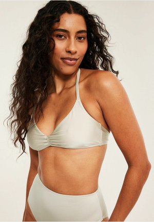 Vrouw met lang krullend haar die een lichtgrijs halterbikinitopje en een bikinibroekje met hoge taille draagt tegen een effen achtergrond.