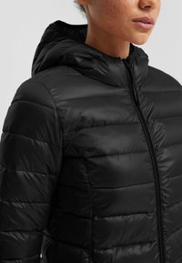 Mustra musta puffer-jakk, millel on lukk ees, kapuuts, sünteetiline õmblus ja horisontaalne õmblus. Sile, läikiv materjal.