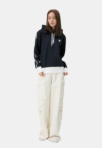 Felpa blu navy con profili e lacci bianchi, abbinata a pantaloni cargo beige con più tasche e cerniere.