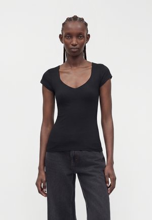 Femme aux cheveux tressés portant un haut noir ajusté à manches courtes en V et un jean gris foncé ample sur un fond clair uni.