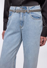Jeans di denim azzurro chiaro con una vestibilità ampia, caratterizzati da borchie argentate e una cintura a fiocco metallico. Tessuto testurizzato con dettagli di cucitura in evidenza.