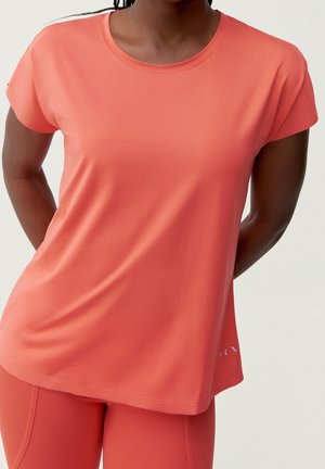 Mujer con camisa deportiva de manga corta color coral y mallas a juego, de pie con las manos detrás de la espalda sobre un fondo liso.