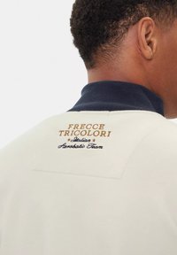 Felpa bianca con colletto blu navy, con ricamati in oro e blu "FRECCE TRICOLORI" e "Squadra Aerea Italiana". Texture liscia.