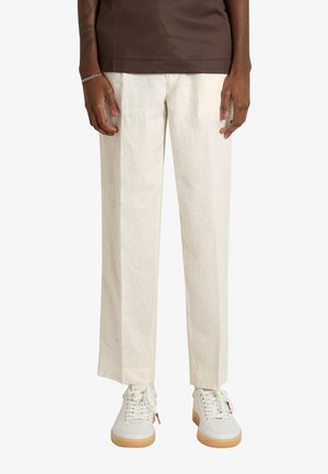 Persona che indossa pantaloni dritti color avorio, sneaker bianche con suole in gomma e una camicia marrone a maniche corte, in piedi su uno sfondo semplice.