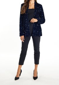 Blazer de lentejuelas azul marino con apertura frontal y mangas largas, combinado con pantalones ajustados negros y tacones de punta negra. Textura de tela suave.