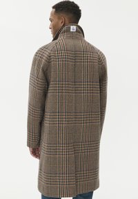 Vīrietis valkā garu brūnu raibumu virsjak, ar augstu apkakli, kas pacelta, atklājot izmēra etiķeti kakla aizmugurē.