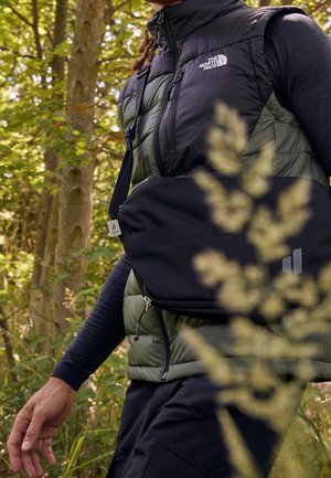 Personne portant une veste North Face noire et verte et un t-shirt noir à manches longues avec un sac bandoulière noir Deuter, marchant dans une forêt.