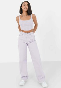 Top corto lilla in tessuto a coste abbinato a jeans larghi a vita alta lilla. Modella che indossa sneakers bianche, in posizione eretta.