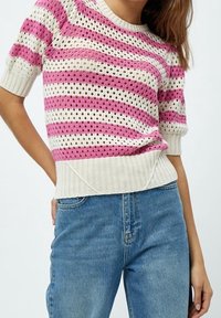 Žena v bílém a růžovém pruhovaném krátkém pleteném svetru s kulatým výstřihem a modrých vysokých džínech.