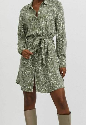 Woman wearing a green long-sleeve, button-up dress with white irregular dot print, belted waist, front slit, and beige knee-high boots.