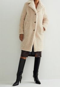 Abrigo beige y esponjoso con cuello de muesca, botones frontales y bolsillos laterales; superpuesto a una falda marrón oscuro y combinado con botas negras hasta la rodilla.