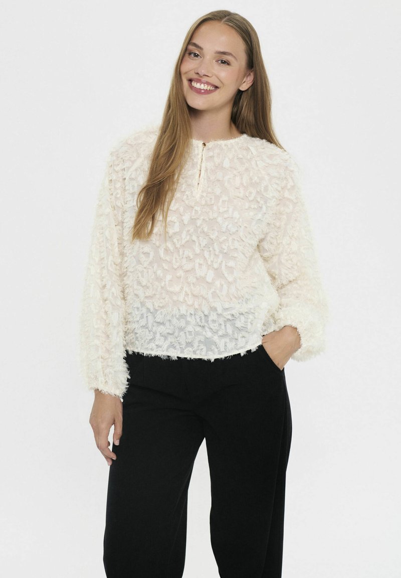 Donna sorridente con capelli lunghi, che indossa una blusa cremosa con texture e pantaloni neri, in piedi con una mano in tasca su uno sfondo bianco.