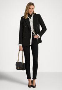 Svart dobbelspent blazer over en hvit skjorte, kombinert med svarte smale bukser og svarte hæler. Bærer en svart håndveske med kjedestropper.