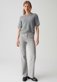 Hellgrauer, kurzärmeliger Pullover aus weichem Material, kombiniert mit hellgrauen Schlagjeans. Das Model trägt schwarze Schuhe und steht in einem neutralen Raum.