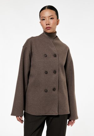 Vrouw met steil zwart haar draagt een bruine dubbelgeknoopte wollen jas over een bijpassende coltrui en zwarte krijtstreepbroek, staand tegen een witte achtergrond.