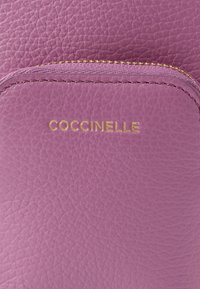 Carteira roxa em couro com acabamento texturizado, nome da marca "Coccinelle" em relevo dourado e fecho de correr com puxador metálico combinando.