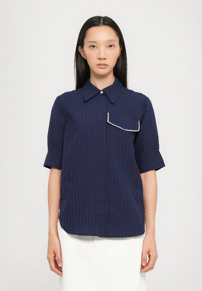 Camicia blu navy a maniche corte con un motivo testurizzato, colletto a punta e un dettaglio di tasca bordato in bianco sul lato sinistro del petto.