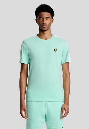 Junger Mann steht, trägt ein hellgrünes kurzärmeliges T-Shirt und passende Shorts mit kleinem gelbem Vogel-Logo auf Brust und Bein.