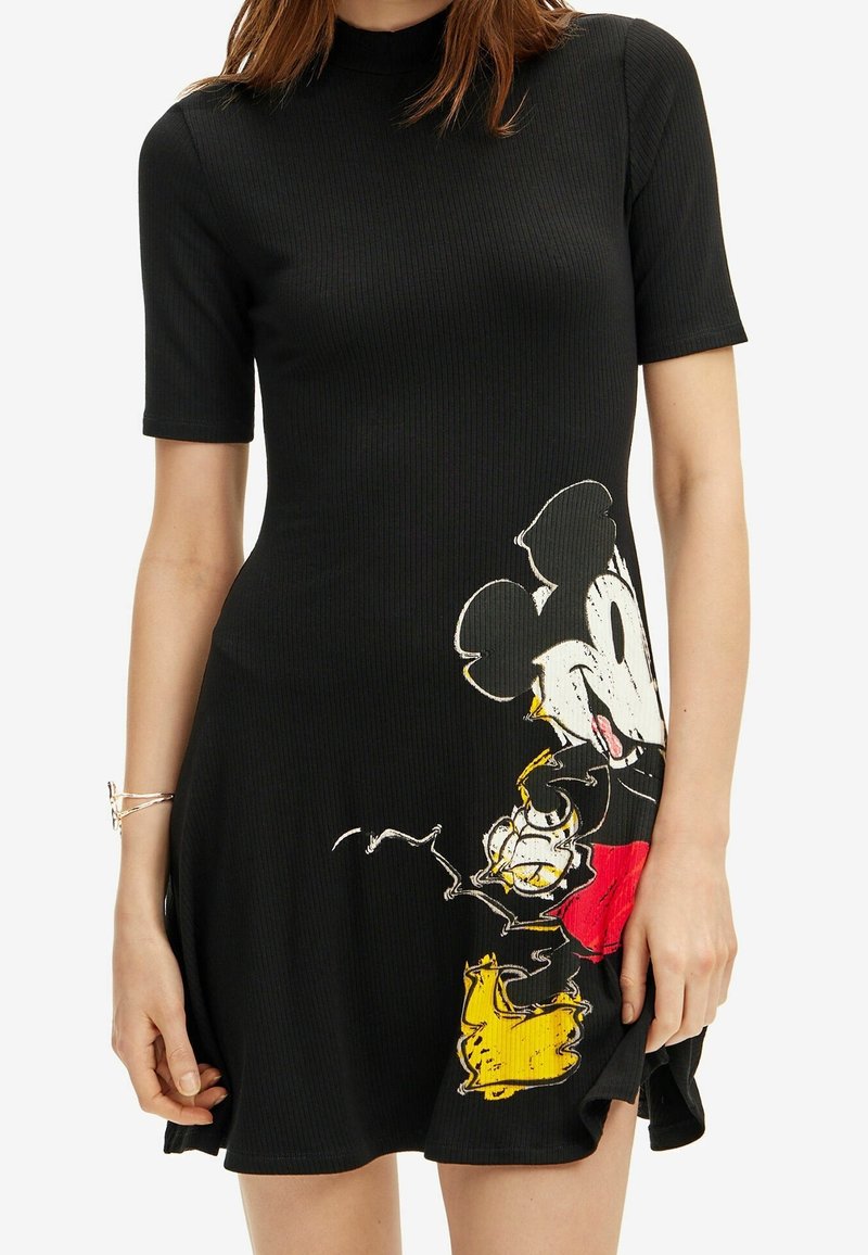 Femme portant une robe noire à manches courtes côtelée avec un grand motif Mickey Mouse usé sur le bas devant.