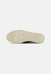 Svarte slip-on sko med overdel i strukturert lerret og beige gummisåle. Har et sikksakkmønster for grep og merkevarelogo på sålen.