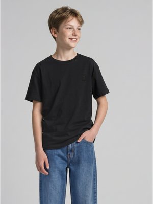 Ragazzo adolescente sorridente con capelli castano chiaro, indossa una t-shirt nera e jeans blu, in piedi con una mano in tasca.