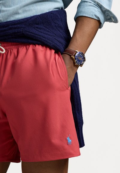 Pantaloni scurți roșii de înot cu un talie elastică, șnur alb și un logo albastru brodat. Un pulover bleumarin este legat în jurul taliei.