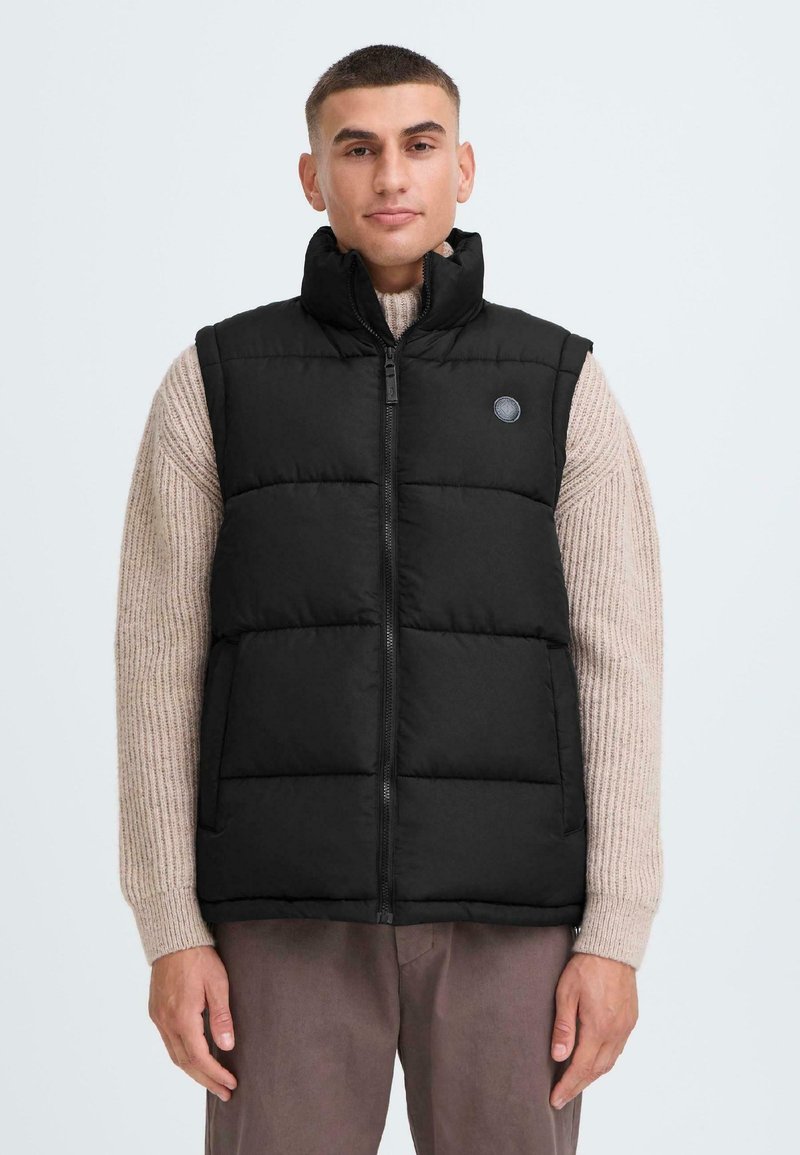 Mustame padded vest kõrge kraega, esipiduri ja logodetailiga. Kantud kerge kudumisega sviitri üle, millel on soonikkoes ja pikad varrukad.