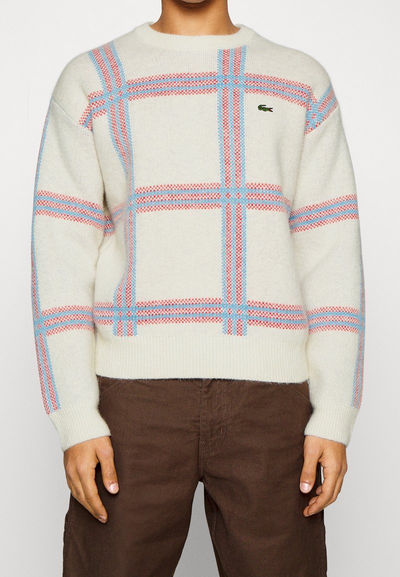 Personne portant un pull crème avec des rayures à carreaux rouges et bleues et un pantalon marron, debout devant un fond uni.