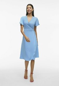 Vaaleansinisiä kietaisumekko, jossa on lyhyet liehutuslahkeet, teksturoitu kangas ja vyötärölle solmittava nauha. Yhdistetty punaisiin soljettukiin sandaaleihin.