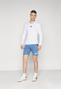Witte, lange mouwen sportshirt met zwart logo, gecombineerd met blauwe shorts met gele strepen en een logo, gedragen met witte sportschoenen.