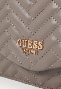 Grå faux læder håndtaske med quiltet mønster, guld "GUESS" logo og syede detaljer. Kompakt design med flaplukning.
