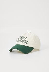 Căciulă de baseball crem cu vizor verde, având brodat textul "OBEY STUDIOS" în verde închis, fabricată dintr-un material durabil.