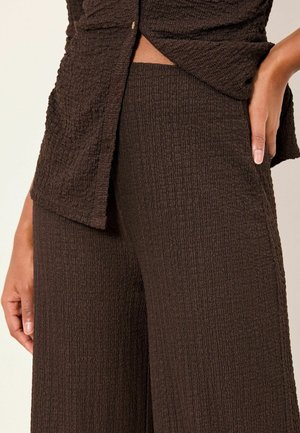 Brun tekstureret top med knapdetalje, kombineret med matchende brune bukser med vide ben. Stoffet har et subtilt krømmønster og en blød finish.