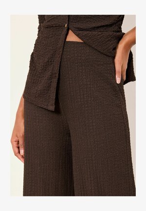 Top marrón texturizado con detalle de botones, combinado con pantalones de pierna ancha a juego en marrón. La tela presenta un sutil patrón arrugado y un acabado suave.