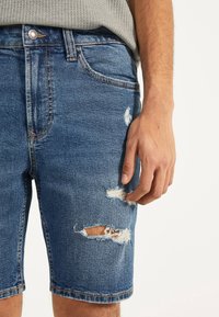 Högra handen vilar på blå denimshorts med slitna och rivna detaljer, som bärs tillsammans med en grå texturerad topp.