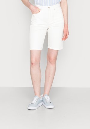 Persona que lleva puestos unos pantalones cortos blancos de talle alto hasta la rodilla y zapatillas gris claro con cordones blancos, de pie frente a un fondo liso.