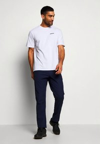 Balti trumpomis rankovėmis marškinėliai su mažyčiu juodu logotipu, derinami su tamsiai mėlynomis kelnėmis ir juodais batais, stovint ant šviesios fono.