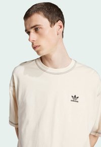 T-shirt di cotone beige con maniche corte. Presenta il logo Adidas nero sul petto e cuciture decorative lungo il collo.
