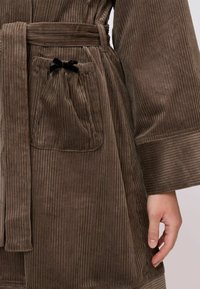 Bársony anyagú köntös gazdag barna színben, függőleges csíkokkal, derékban megkötős, zsebén fekete szalag díszítéssel. Puha tapintású és széles ujjú.